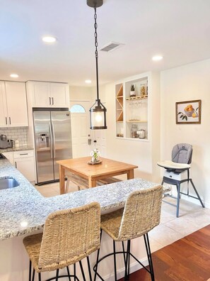 A modern kitchen with white cabinets, a stainless steel refrigerator, a wooden dining table, wicker chairs, and a baby high chair. - Beautiful 3 BR Home by the Water- Family Friendly (Weymouth)
