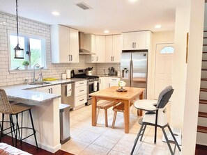 A modern kitchen with white cabinets, a stainless steel refrigerator, a wooden dining table, and a bar stool. - Beautiful 3 BR Home by the Water- Family Friendly (Weymouth)
