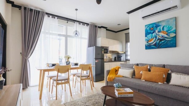 Ein modernes Wohnzimmer mit grauem Sofa, Esstisch aus Holz und Stühlen. Es gibt eine Küchenzeile mit Schränken und einem Kühlschrank.
