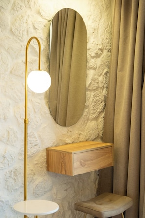 A bathroom with a stone wall, a wooden vanity, a round mirror, a gold floor lamp, and a white stool.