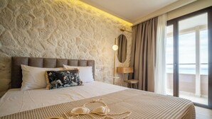 A bedroom with a stone wall, a bed with white linens, a nightstand, and a view of the sea.