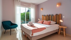 Un dormitorio con cama, una silla, mesita de noche con lámpara y vistas al exterior con vegetación.