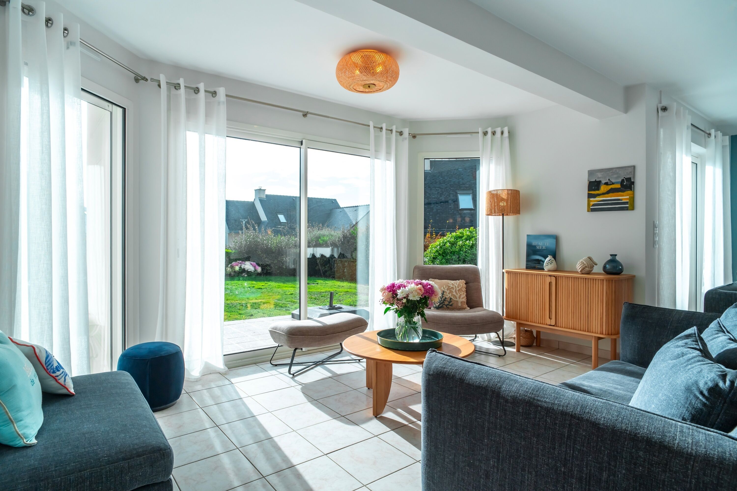 Sala de estar moderna com um sofá, poltrona e mesa de centro com flores. Há um armário de madeira e uma luminária. O ambiente possui janelas amplas com cortinas e vista para o exterior.