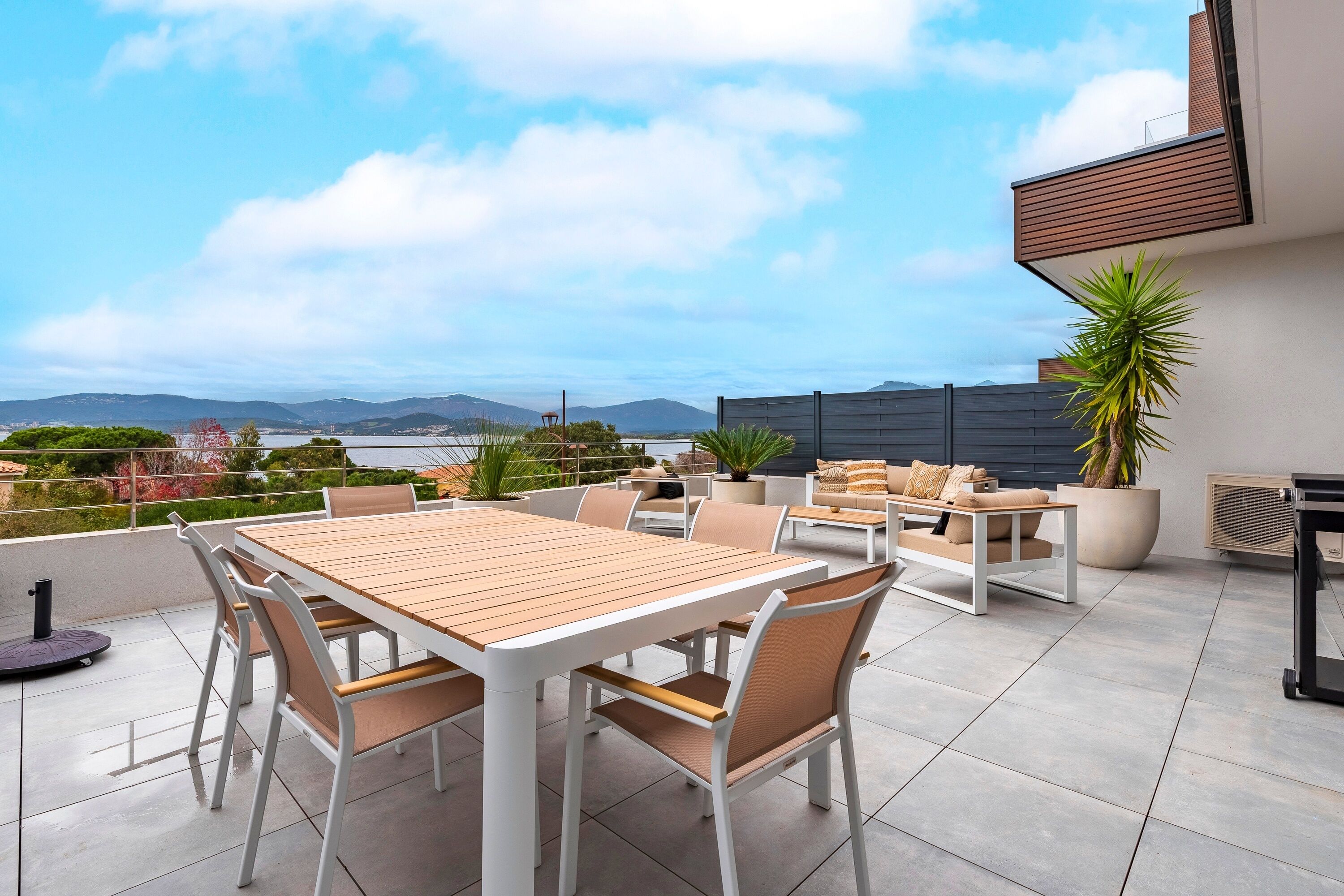 A rooftop terrace with a wooden table and chairs, a potted plant, and a view of the sea and mountains.