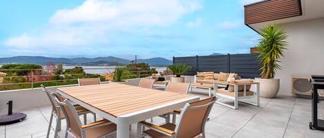 A rooftop terrace with a wooden table and chairs, a potted plant, and a view of the sea and mountains.