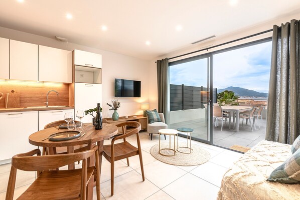 A modern kitchen with wooden cabinets, a dining area with a round table and chairs, a sofa, and a large sliding glass door leading to a balcony with a view of a cityscape and mountains. - A Terrazzo di Calypso - Grande Terrasse (Grosseto-Prugna)