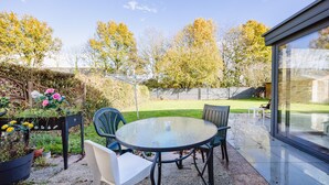Une terrasse dotée d’une table ronde en verre et de quatre chaises, entourée d’un jardin où poussent différentes plantes et fleurs.