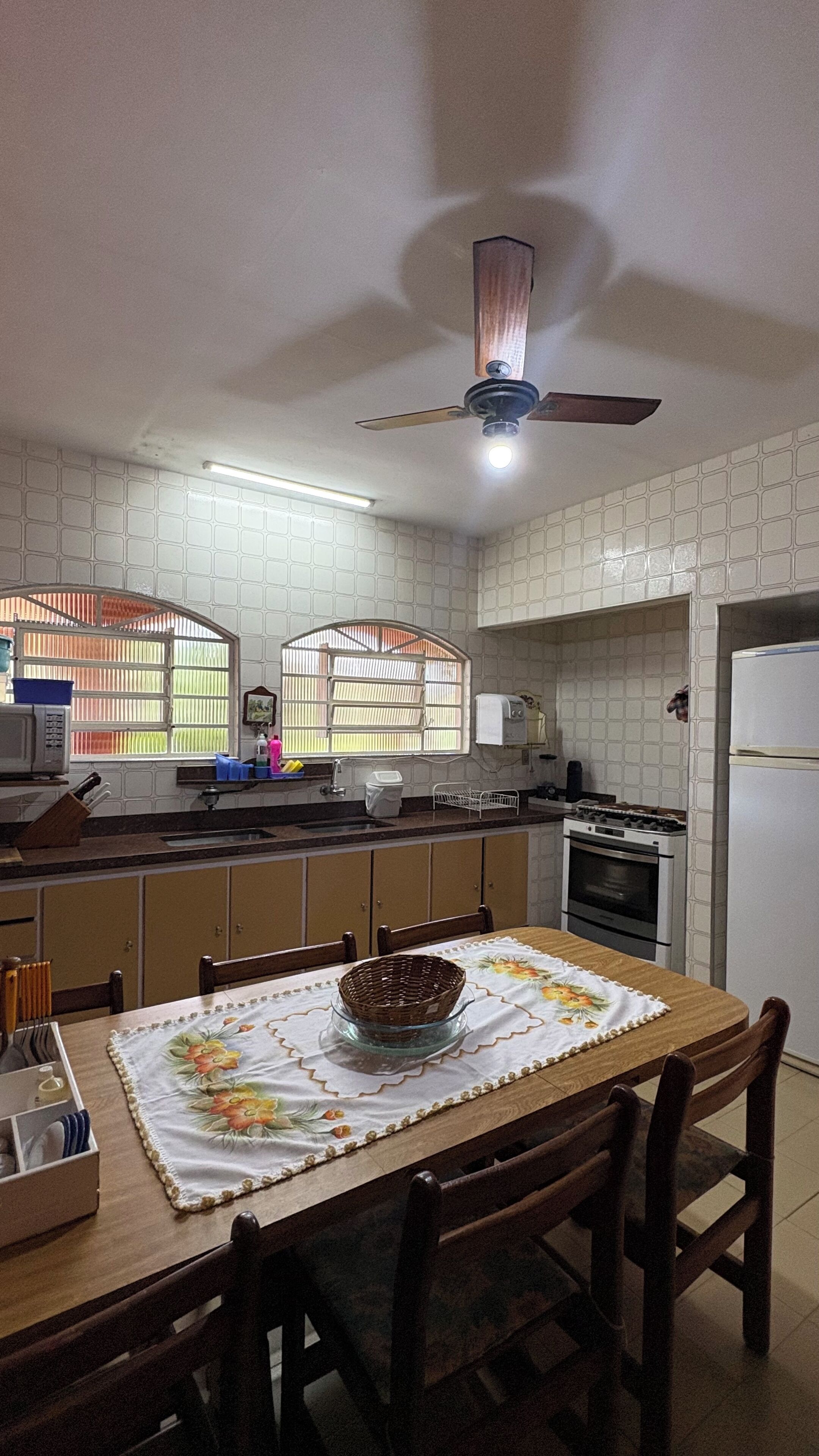 Una cocina con mesa, sillas, ventilador de techo, refrigerador y ventana con persianas.