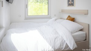 Ein Schlafzimmer mit Bett, Fenster, einem an der Wand montierten Fernseher und einem gerahmten Bild.