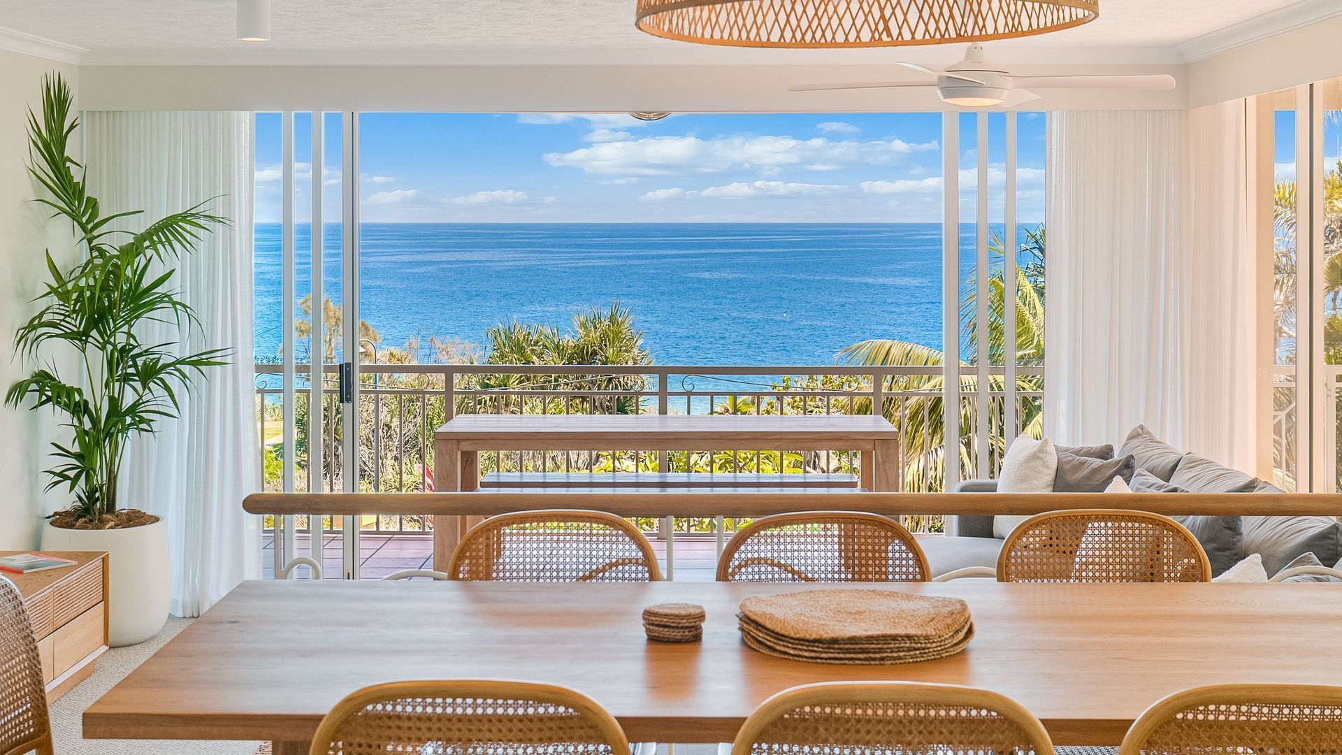 A dining area with a view of the ocean, featuring a wooden table, wicker chairs, and a ceiling fan.