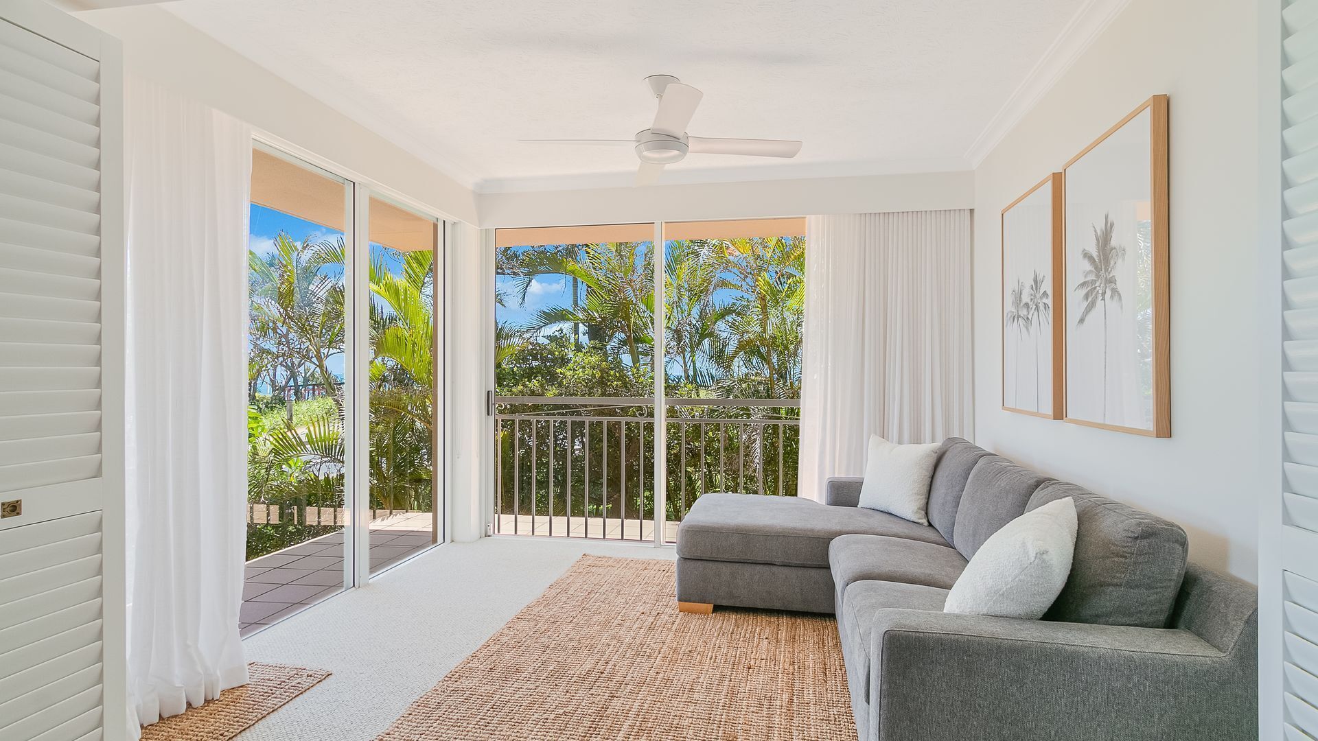 A spacious living room with a grey sofa, a ceiling fan, and a view of lush greenery through large sliding glass doors.