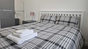 A neatly made bed with a checkered bedspread, a bedside lamp, and a stack of folded towels. - Stylish 2 Bed Flat Near Airpot with free parking (Edinburgh)