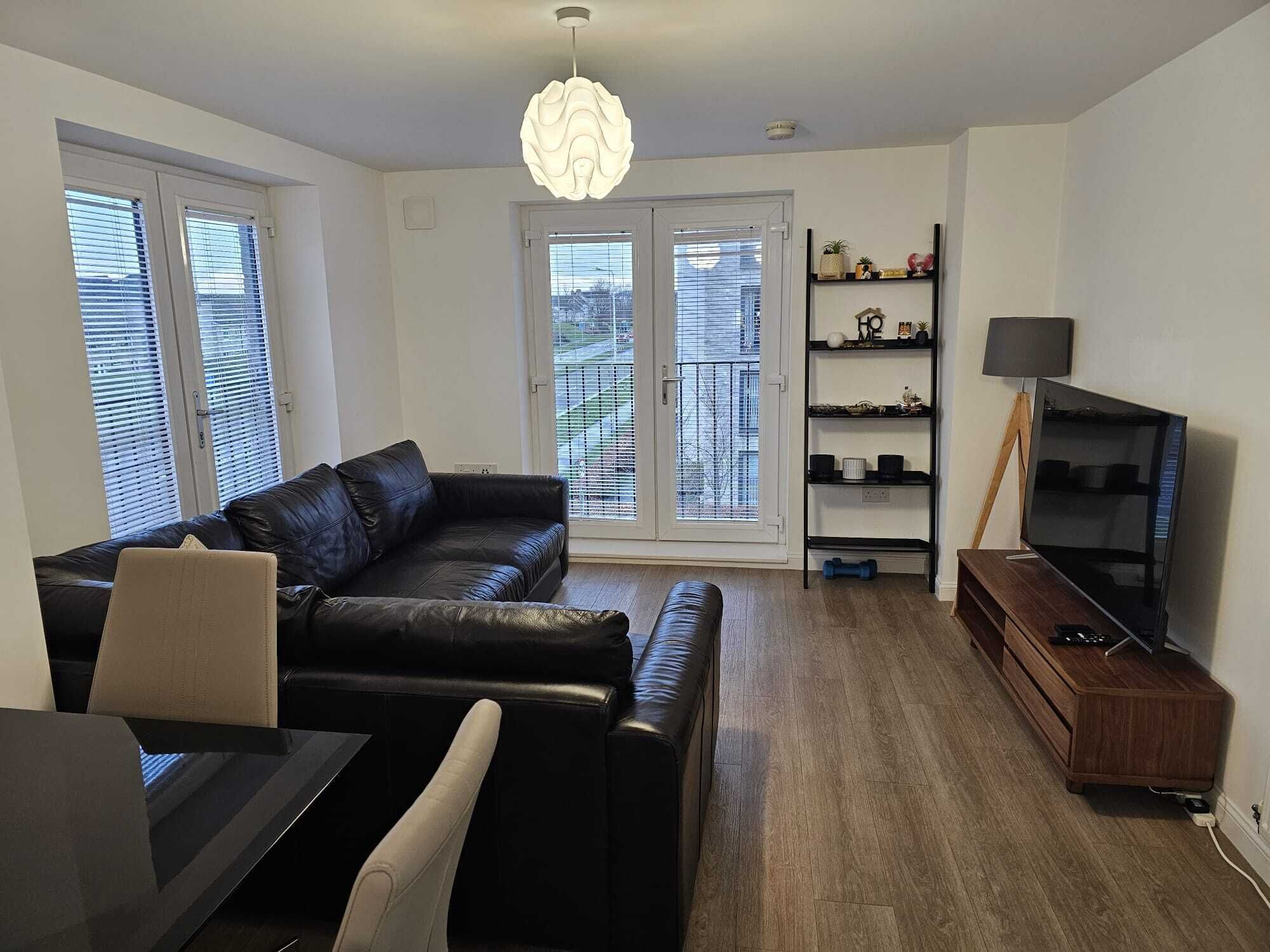 Una sala de estar moderna con un sofá de cuero negro, una mesa de comedor con sillas, un televisor sobre un mueble de madera y una estantería.