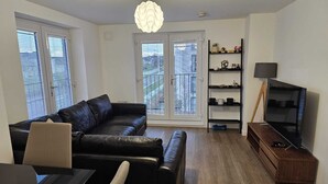A modern living room with a black leather sofa, a dining table with chairs, a TV on a wooden stand, and a bookshelf. - Stylish 2 Bed Flat Near Airpot with free parking (Edinburgh)