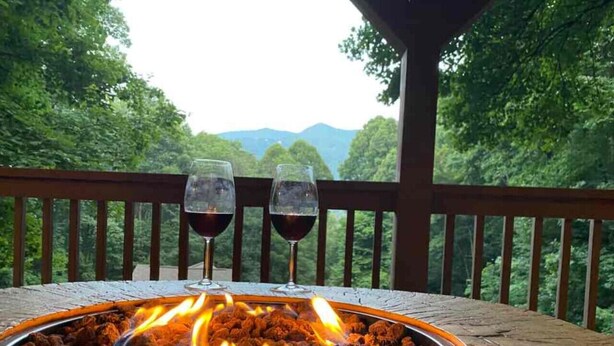 Uma mesa com lareira, com dois copos de vinho tinto, cercada por um deck de madeira e vista para uma cadeia montanhosa arborizada.