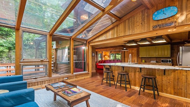 A spacious kitchen with a wooden interior, a bar area with stools, and a large window overlooking a forested area. - Sasha’s Hidden Creek Cabin-Magical Views, 12 Acres (Maggie Valley)