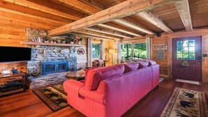 A cozy cabin interior with a stone fireplace, wooden beams, a red sofa, and a television. - Sasha’s Hidden Creek Cabin-Magical Views, 12 Acres (Maggie Valley)