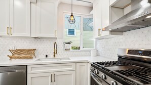 Uma cozinha moderna com armários brancos, lava-louças de aço inoxidável, fogão a gás e uma janela com vista para o verde.