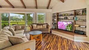 A spacious living room with a large sofa, a coffee table, and a TV on a wooden stand. There are large windows with a view of a green landscape.