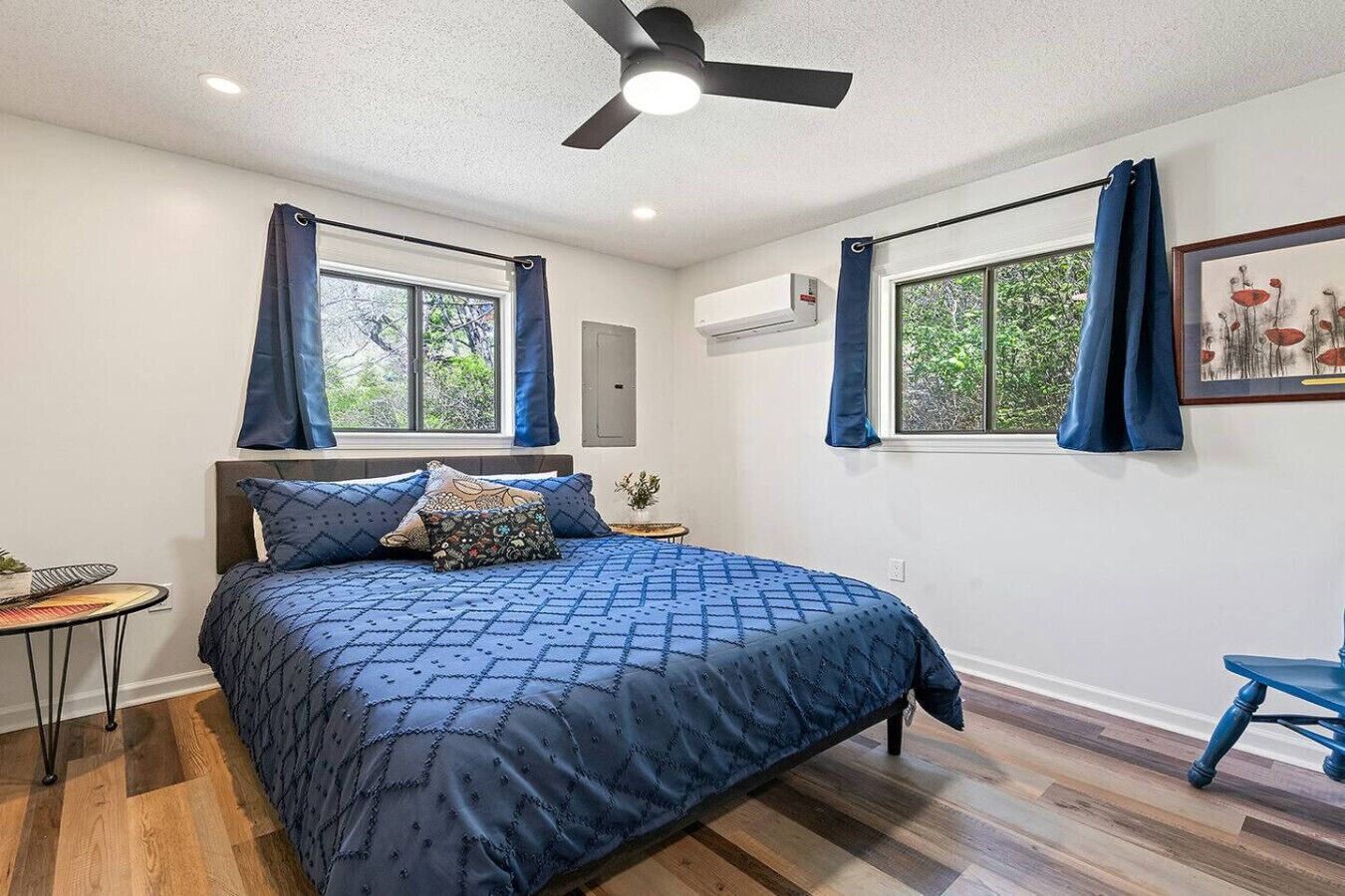 Un dormitorio con cama, ventilador de techo, dos ventanas con cortinas, una mesita de noche con una planta y un cuadro en la pared.