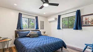 Una camera da letto con un letto, una ventola a soffitto, due finestre con tende, un tavolino con una pianta e un quadro appeso al muro.