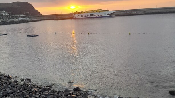 Ein Sonnenuntergang über ruhigem Wasser mit einer Fähre namens 'FRED. OLSEN Express' und einer felsigen Küstenlinie.