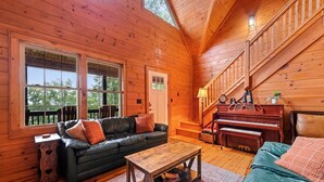 A cozy cabin interior with a wooden floor, a black leather sofa, a wooden coffee table, a piano, and a staircase. - Falcon’s Nest-Spacious w/Spectacular Views!Hot Tub (Maggie Valley)