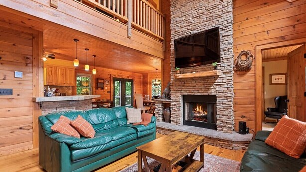 A cozy cabin interior with a stone fireplace, wooden floors, a green leather sofa, and a wooden dining table. - Falcon’s Nest-Spacious w/Spectacular Views!Hot Tub (Maggie Valley)