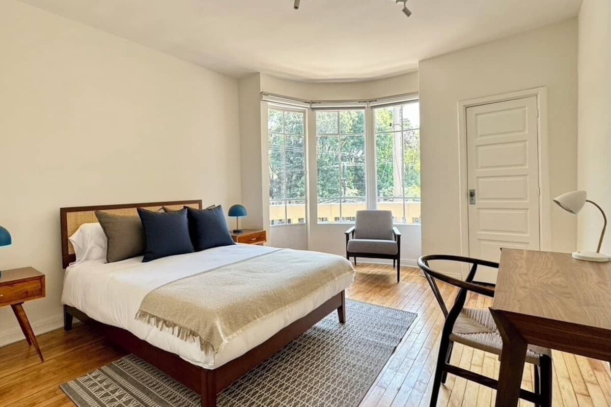 Quarto com cama, mesas de cabeceira, uma cadeira, escrivaninha e uma janela grande.