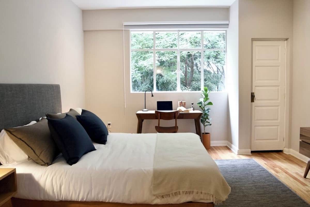Quarto com cama, escrivaninha, cadeira, planta em vaso e janela com vista para árvores.