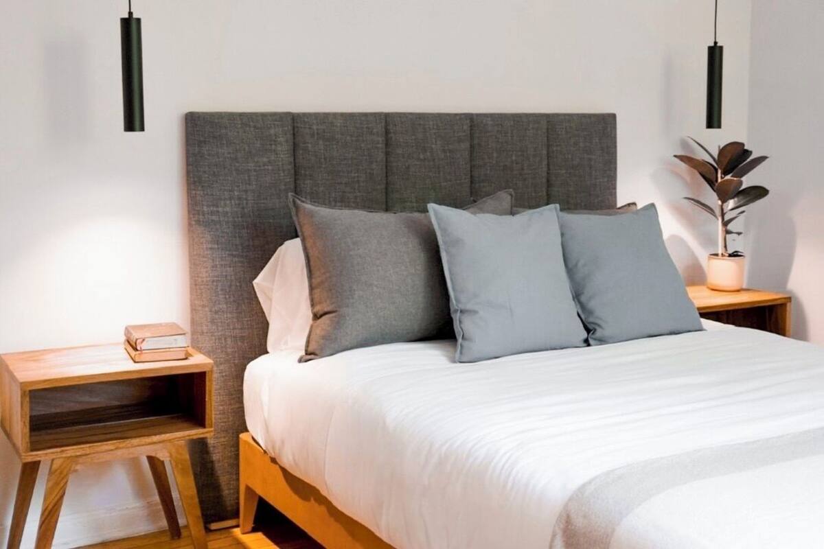Quarto com cabeceira estofada cinza, mesa de cabeceira de madeira com abajur e planta ao lado da cama.