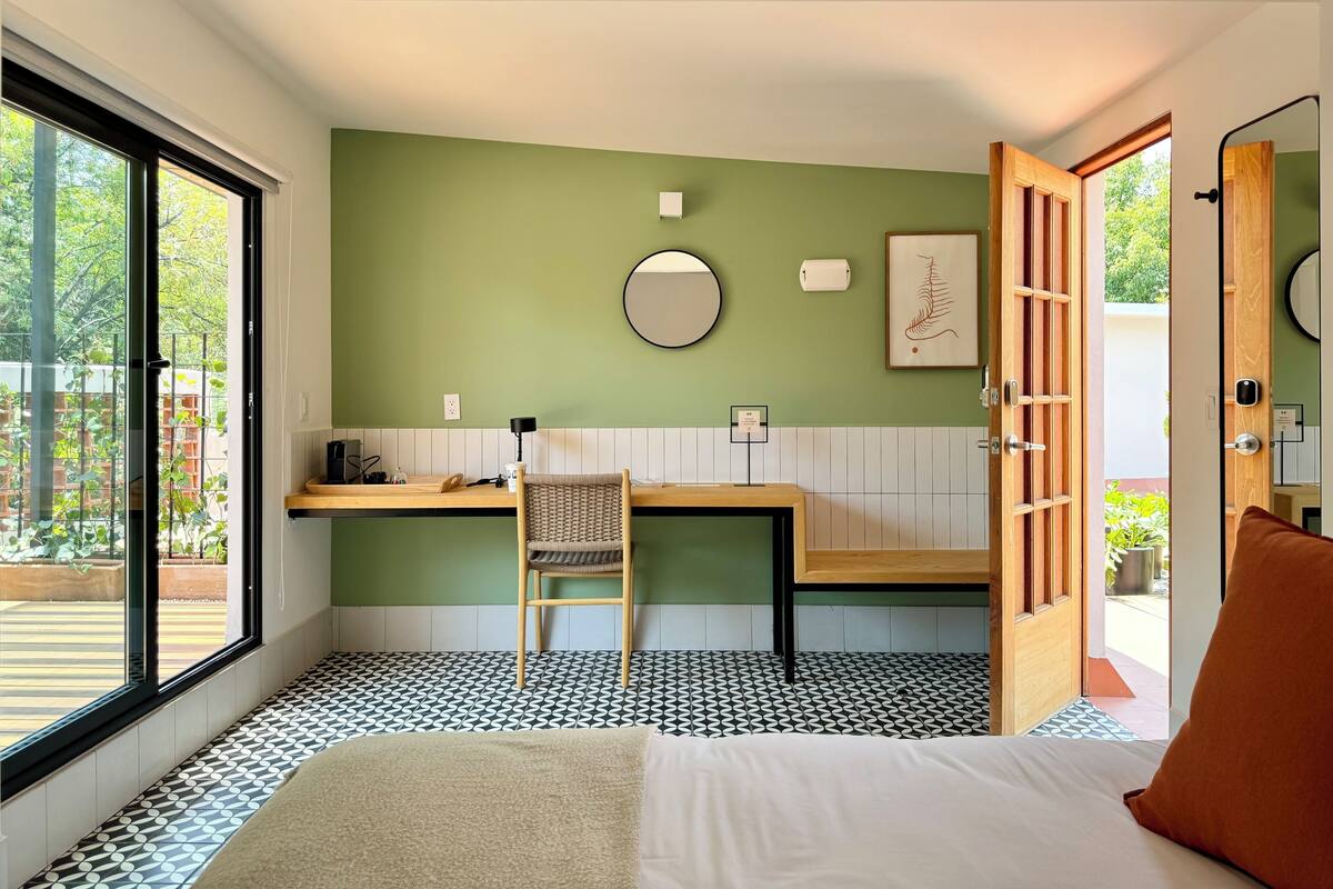 Quarto com cama, escrivaninha, cadeira, espelho e vista para o exterior.