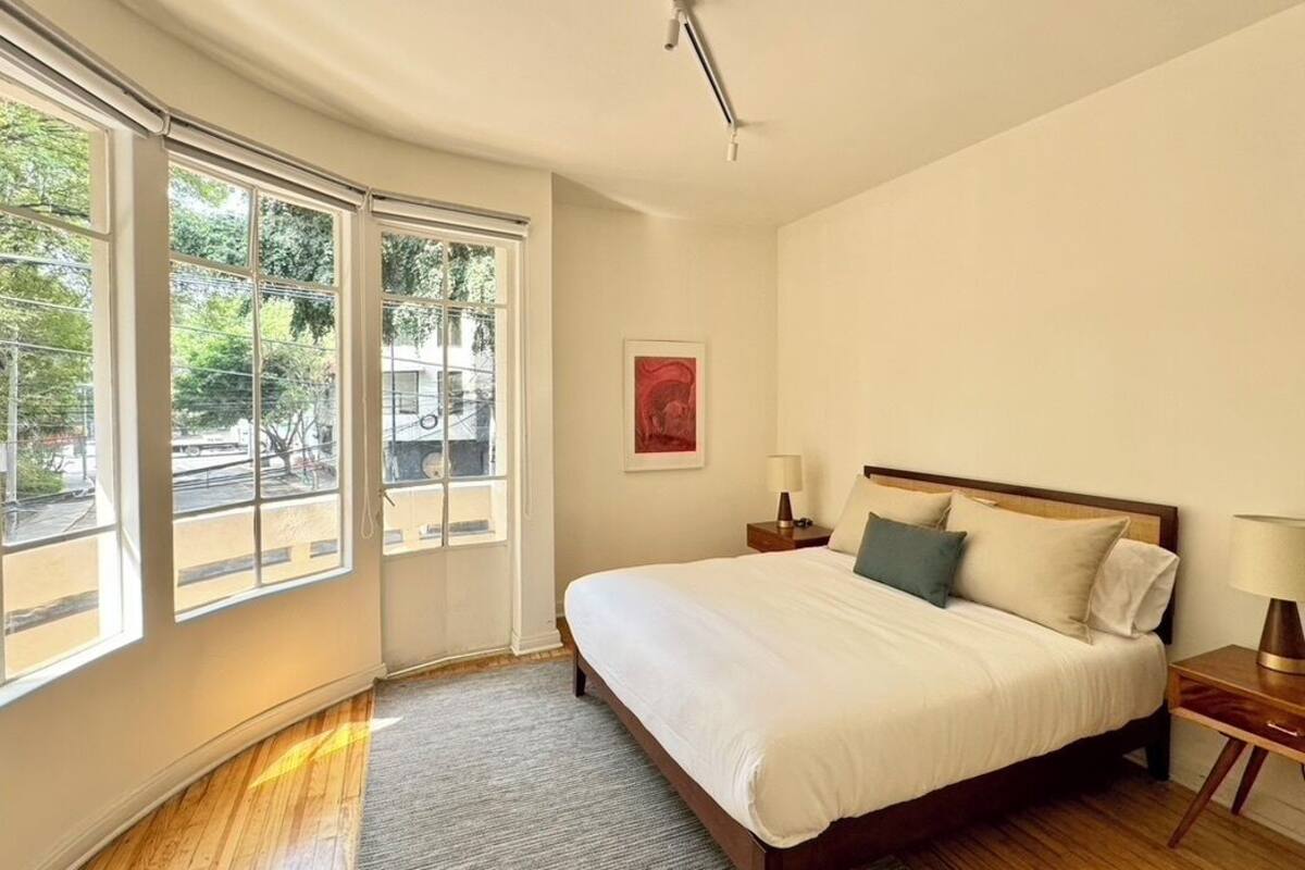 Quarto com uma cama grande, mesa de cabeceira, luminária e janela com vista para uma linha de trem.