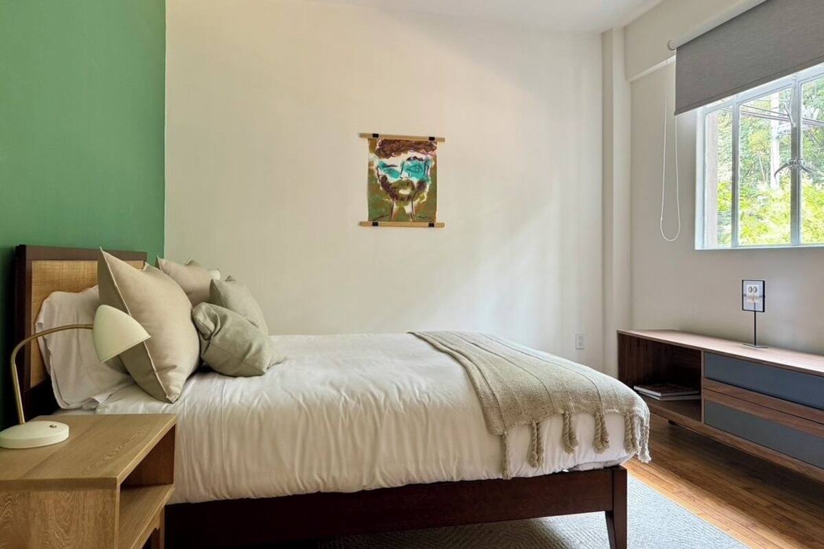 Quarto com cama, mesa de cabeceira, abajur e janela com vista para o verde.