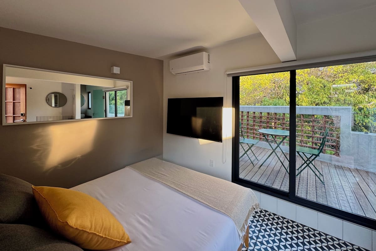 Um quarto com uma cama, um espelho grande, uma TV fixada na parede e vista para um deck exterior com uma mesa e cadeiras.