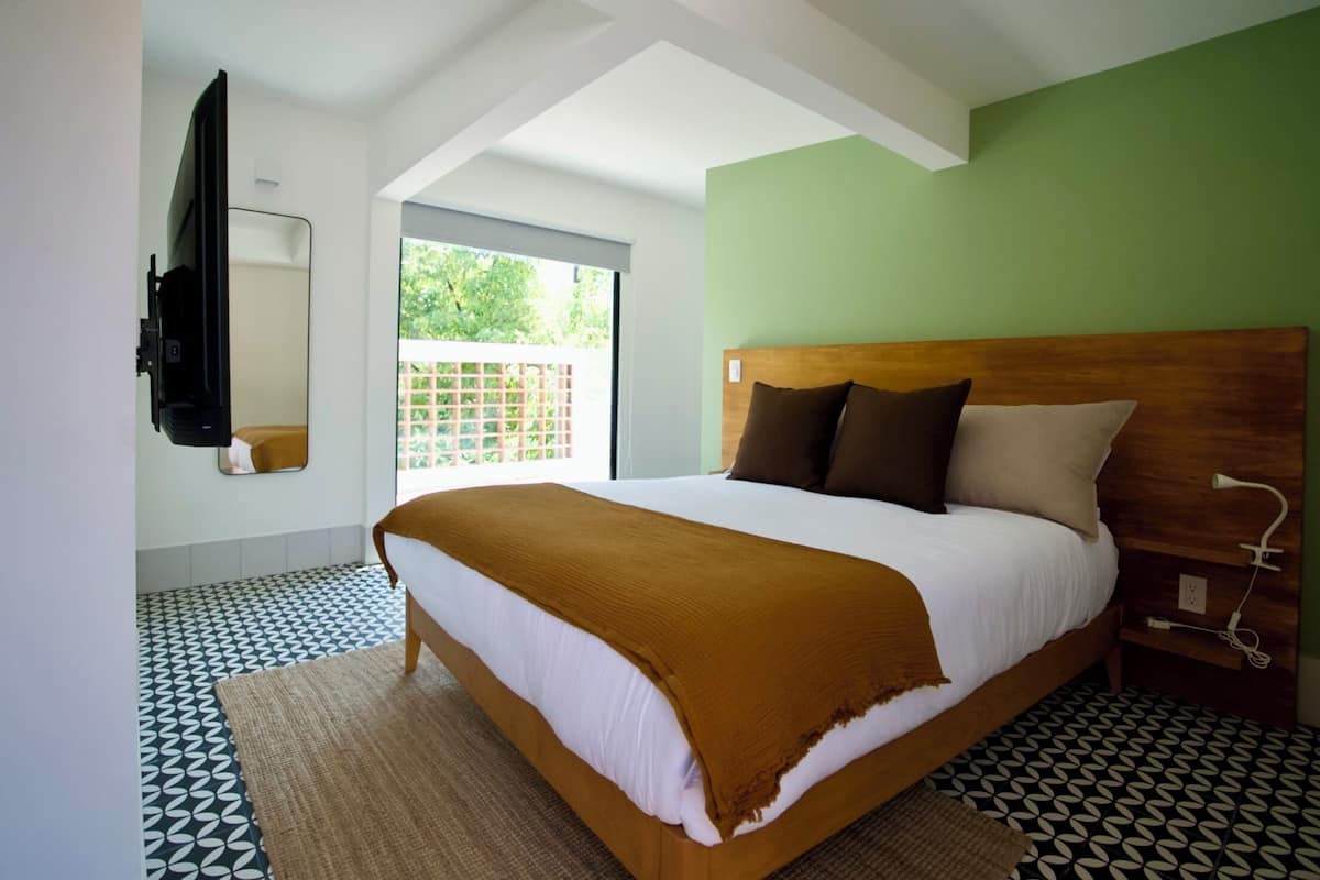 Quarto com cama grande, cabeceira de madeira, espelho, televisão e janela com vista para o verde.