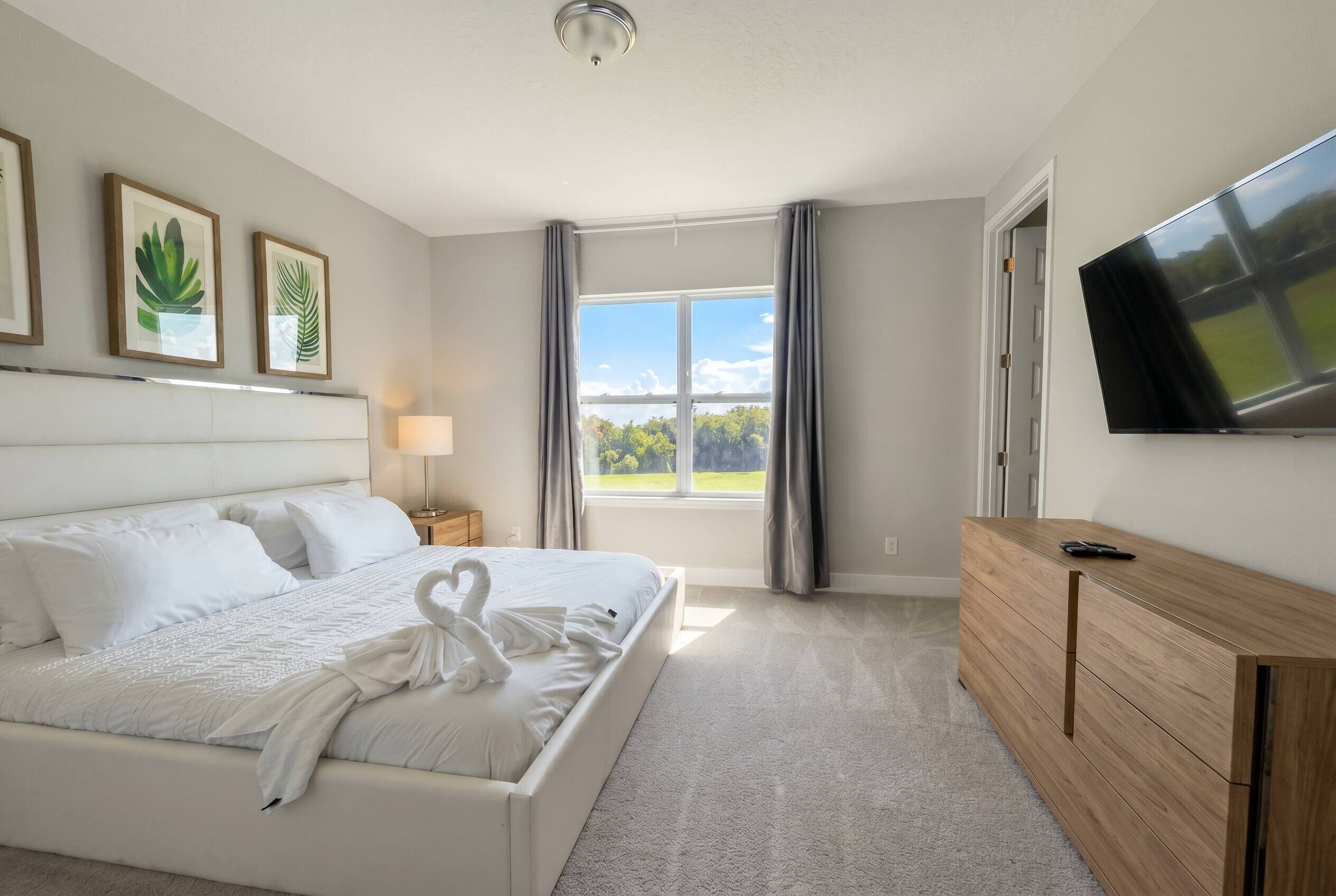 Una camera d'albergo con un letto, una televisione, un comò, una finestra con tende e quadri appesi al muro.