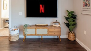 Een moderne woonkamer met een flatscreen televisie waarop het Netflix-logo staat, een houten salontafel met decoratieve objecten, een rieten mand en een grote groene plant.