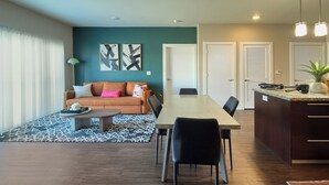 Een moderne woonkamer met een bank, salontafel en een eethoek met stoelen en een tafel. Er is een keuken met een aanrecht en kasten.
