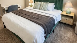 Een net opgemaakt bed met een groen hoofdeinde, witte beddengoed en een bruine plaid. Er staat een bureau met een stoel, een lamp op het nachtkastje en een klein lade.