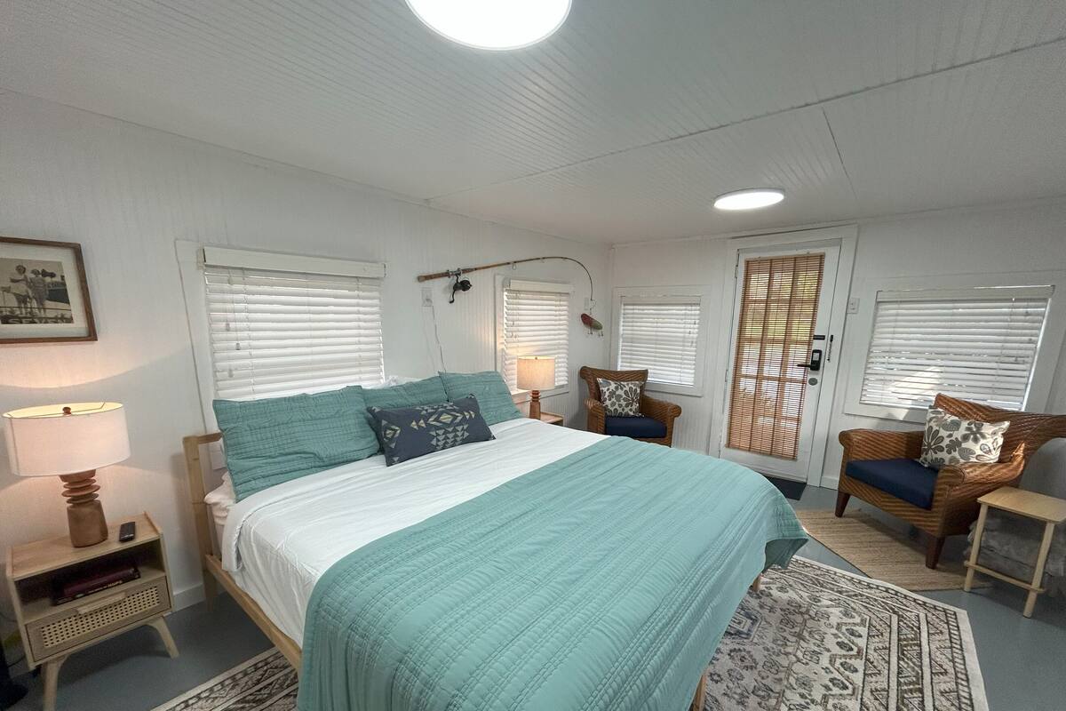 Un dormitorio con cama, una silla, una mesita de noche, una lámpara y una ventana con persianas.