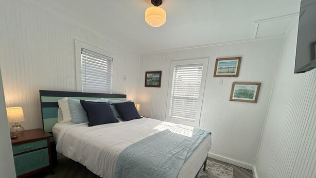 A bedroom with a bed, nightstands, a lamp, a window with blinds, and a wall-mounted TV. - Old Florida Fish Camp (Cedar Key)