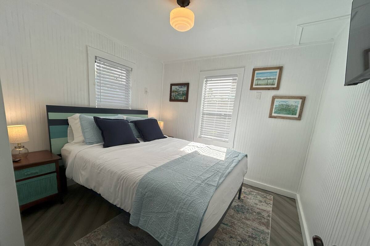 Un dormitorio con cama, mesitas de noche, lámpara, ventana con persianas y televisor instalado en la pared.