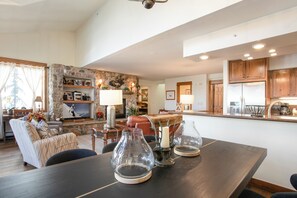A spacious living area with a stone accent wall, a dining table with clear glassware, and a kitchen with wooden cabinets. - Beautiful 4 Bedroom Condo with pool and hot tub access. 150 yards to Gondola (Vail)