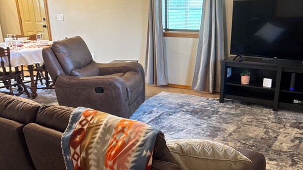 Una sala de estar con un sofá de cuero marrón, un sillón reclinable marrón, un televisor sobre un mueble, una mesa de comedor con sillas y una ventana con cortinas.