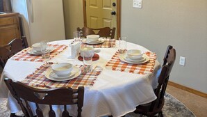Un comedor con una mesa redonda dispuesta para cuatro personas, rodeada de sillas de madera. Un mantel blanco cubre la mesa, y hay platos, tazones y vasos sobre ella. Se ve un refrigerador al fondo, y una puerta en el centro de la pared.