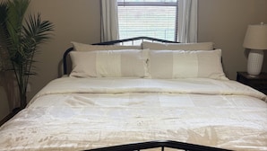Una cama bien tendida con estructura metálica, una ventana con persianas, una lámpara de noche y una planta en maceta.
