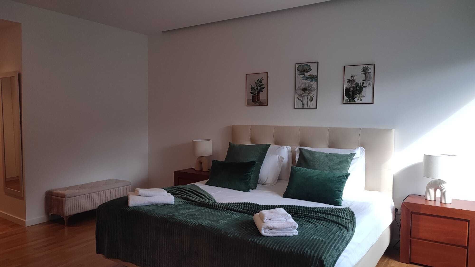 Une chambre d’hôtel avec un grand lit, deux tables de chevet, un banc et trois reproductions encadrées de motifs botaniques accrochées au mur.