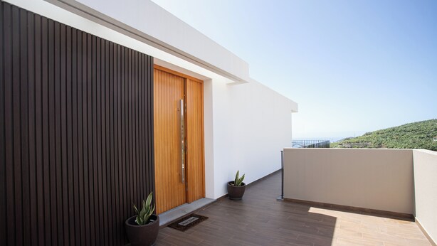 A modern building with a wooden door, potted plants, and a balcony with a view of a green hill. - Dori’s Modern Home w/Pool & Sea & Mountain Views — 15 Min to City Center (Funchal)