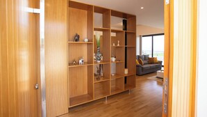 A modern living room with a wooden bookshelf, a grey sofa, and a large window. - Dori’s Modern Home w/Pool & Sea & Mountain Views — 15 Min to City Center (Funchal)